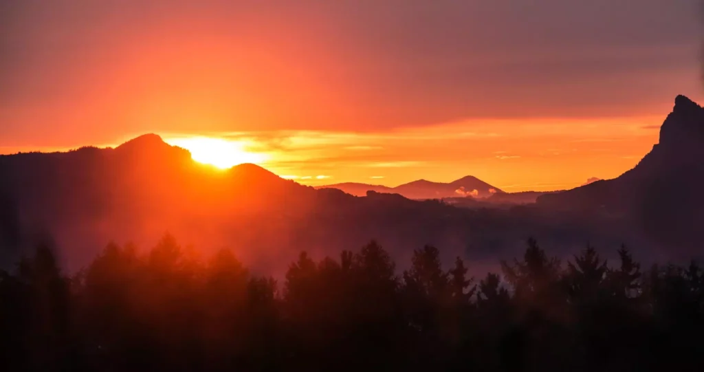 A breathtaking sunset over the mountains, casting a golden-orange glow across the sky and silhouetting the forested landscape below.