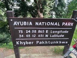 A wooden signboard displaying the coordinates of Ayubia National Park in Khyber Pakhtunkhwa, Pakistan. The sign provides the park's longitude (73° 24' 58.865" E) and latitude (34° 03' 12.031" N), surrounded by lush green trees in the background.