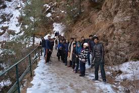 A group of tourists walking along a snow-covered trail in Nathia Gali, Pakistan. The pathway is surrounded by rocky hills and pine trees, creating a scenic winter hiking experience.