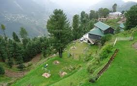 A scenic view of Nathia Gali, Pakistan, featuring lush green hills, tall pine trees, and a beautiful cottage with a green rooftop overlooking the valley. The misty mountains in the background add to the serene and peaceful atmosphere.